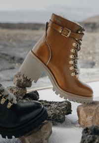 Botas marrones de tacón alto con suela gruesa, que presentan herrajes metálicos, detalles de encaje y un acabado de cuero suave. Bota negra parcialmente visible.
