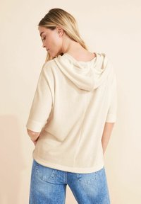 Kremowy wełniany hoodie o luźnym kroju, z krótkimi rękawami i teksturowanym wykończeniem. Posiada szew z tyłu oraz kaptur ze sznurkiem.