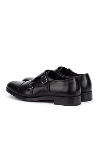 Zapatos de vestir de charol negro con doble hebilla y plateada, tacón bajo y punta redonda, mostrados desde un ángulo lateral y trasero.