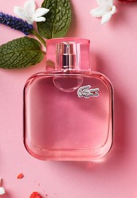 Flacon de parfum carré transparent rose avec un pulvérisateur argenté, entouré de fleurs blanches, de boutons violets et de feuilles vertes sur fond rose.