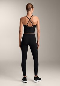 Černý sportovní crop top s kříženými ramínky, kombinovaný s černými legínami s vysokým pasem a černými sportovními botami na neutrálním pozadí.