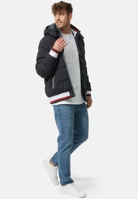 Black puffer jacket with a hood, contrasting red and white ribbed cuffs and hem, zippered front, worn over a white shirt and blue jeans.