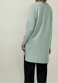 Ljusgrön lång cardigan med avslappnad passform, sidoklyftor och ribbad nederkant. Mjuk textur, matchad med mörka byxor. Minimalistisk design.