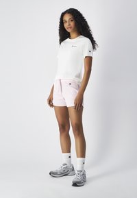 Weißes Kurzarm-T-Shirt mit schwarzem Logo, kombiniert mit hellrosa Shorts, grauen Sportschuhen und weißen Socken mit Logo-Akzenten.