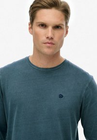 Heren T-shirt met lange mouwen in een donkere teal kleur, gemaakt van zachte stof, met een ronde hals en een klein geborduurd logo op de borst.