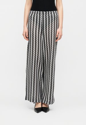 Szerokie spodnie w czarno-białe zygzakowate paski, noszone z czarnymi, spiczastymi butami i czarną bluzką, stojące na jednolitym tle.