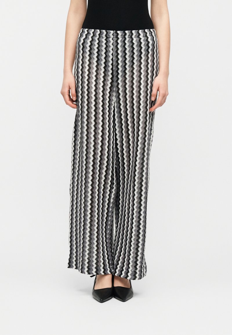 Μαύρο και άσπρο παντελόνι με φαρδιά πόδια και ζικ-ζακ ρίγες, φορεμένο με μαύρα μυτερά παπούτσια και μαύρο τοπ, στέκεται μπροστά σε απλό φόντο.