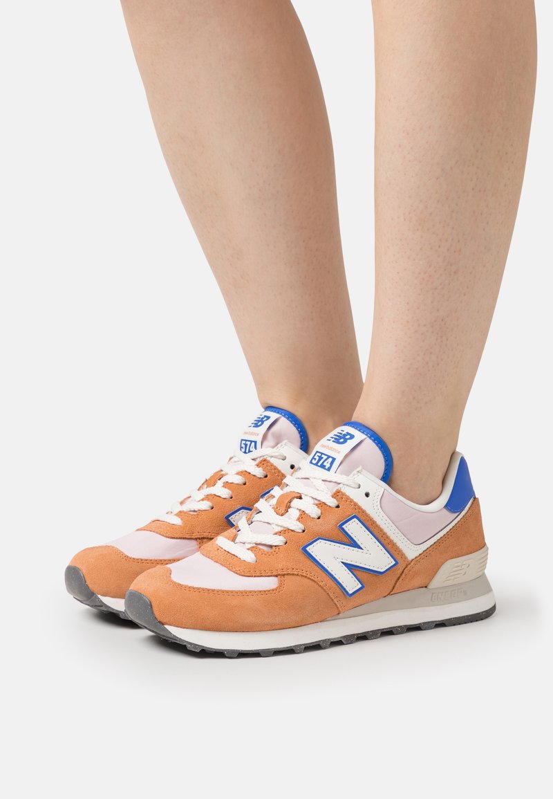 Jambes portant des baskets New Balance 574 orange, blanches et bleues avec des lacets blancs, debout sur une surface blanche.