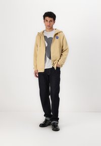 Beige hettegenser med snor, grafisk t-skjorte med et stort design, mørkeblå jeans og svarte sko. Myk stoff og ledig passform.