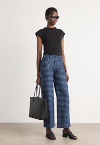 Schwarzes, eng anliegendes T-Shirt, blaue weite Hose mit DKNY-Logo-Muster, schwarze Sandalen mit offenen Zehen und eine schwarze Tragetasche. Glatte Textur, lässiger Stil.
