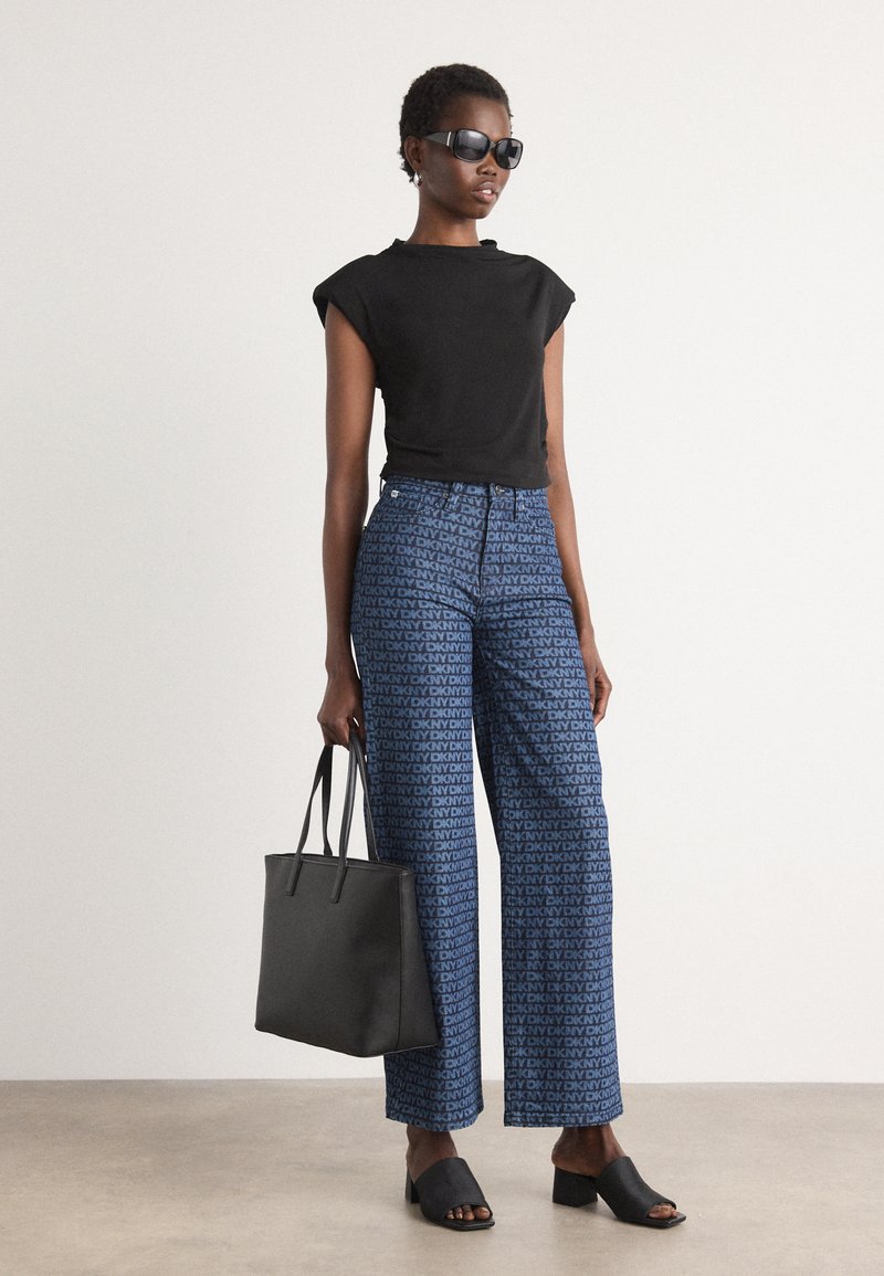 Camiseta negra ajustada, pantalones de pierna ancha azules con patrón de logo de DKNY, sandalias negras de tacón abierto y un bolso tote negro. Textura suave, estilo casual.