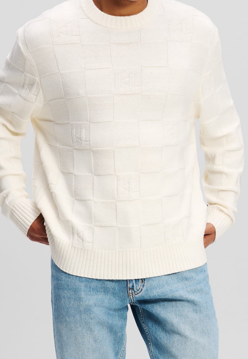 Homme portant un pull en maille blanc texturé avec des motifs à carreaux subtils et un jean bleu clair, debout les mains dans les poches.