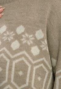 Beige gebreide trui met een wit geometrisch patroon van hexagons en sneeuwvlokken over de borst. Zachte textuur met een ontspannen pasvorm.