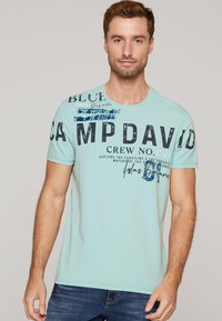 Jasnoniebieska koszulka bawełniana z dużymi czarnymi literami, grafikami i tekstowymi akcentami. Krótkie rękawy i okrągły dekolt.