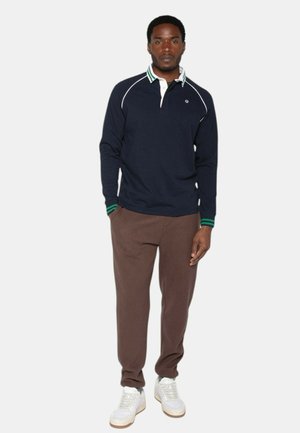 Homme debout, portant un polo à manches longues bleu marine avec col et poignets rayés verts, pantalon marron et baskets blanches, sur fond uni.