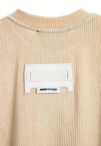 Tela de corduroy beige con canalones verticales, que presenta una etiqueta de cuero blanco con texto en relieve y una etiqueta de logo negra debajo.