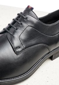 Chaussure en cuir noir avec une texture lisse, un design à lacets, des détails de couture subtils et une accentuation colorée au talon.