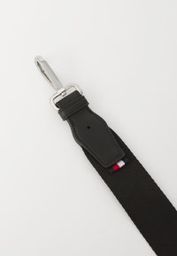 Black elastic strap with a leather attachment, silver metal hardware, and a small tricolor stripe detail near the base.