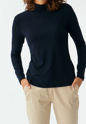 Kobieta ubrana w czarną bluzkę z długim rękawem i golfem oraz beżowe spodnie, z rękami w kieszeniach, na tle jednolitego jasnego tła.