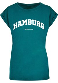 Tyrkysové tričko s krátkým rukávem vyrobené z bavlny, s nápisem "HAMBURG" ve velkém bílém obloukovém písmu a "MERCH•DE" v menším textu pod ním.