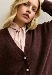 Brun stickad kavaj med V-ringning och tre guldfärgade knappar, över en ljusrosa blus med en knyten krage.