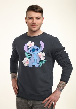 Man met kort bruin haar, tatoeages op de onderarmen, gekleed in een zwarte trui met het personage Stitch en een bloemmotief, staand met handen in de zij.