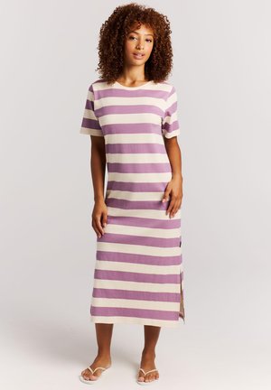 Vrouw met krullend haar, gekleed in een midi-jurk met brede paarse en witte strepen en witte slippers, staand tegen een effen lichte achtergrond.