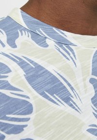 Närbild av en blå och vit mönstrad skjortkrage på mörk hud, med abstrakta bladliknande former och fina horisontella linjer.