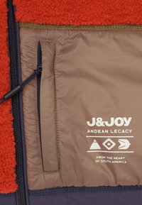 Rood fleece met een bruin nylon voering heeft een ritszak en witte merktekst. Inclusief textuurmaterialen en gekleurde stiksels.