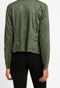 Persona vistiendo una chaqueta de ante verde oliva ajustada y pantalones negros, vista desde la espalda contra un fondo liso.