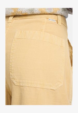 Pantalones cortos de tela texturizada amarilla con un bolsillo trasero y una pequeña etiqueta de la marca en la cintura. La vista lateral muestra la ligereza del material.
