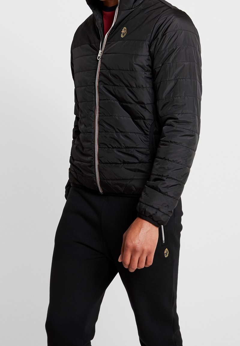 Homme portant une veste matelassée noire zippée et un pantalon noir avec un emblème doré, debout devant un fond clair uni.