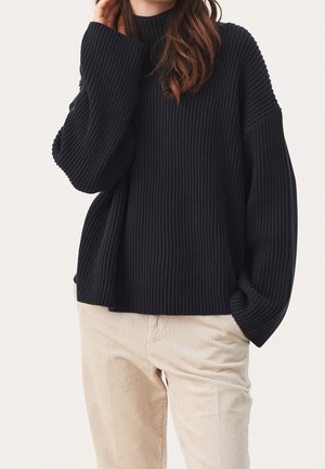 Femme portant un pull oversize noir côtelé à manches ballon et un pantalon en velours côtelé beige, main près du visage, sur fond uni.