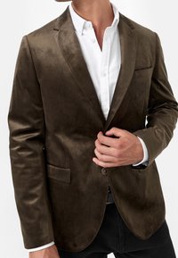 Mørkebrun blazer i et glat stof, med enkelt knaplukning, notch-lapel og en brystlomme, parret med en hvid skjorte.