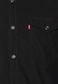 Veste en velours côtelé noir avec boutons-pression argentés et poche poitrine ornée d'une étiquette rouge Levi's. Tissu texturé à côtes verticales.