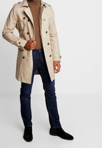 Beiger Trenchcoat mit braunen Knöpfen, doppelt knopfbar, mit Seitentaschen, kombiniert mit dunkelblauen Jeans und schwarzen Wildleder-Stiefeletten.