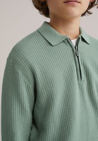 Jeune personne portant un pull en maille texturée vert clair avec un col zippé par-dessus une chemise blanche, fond neutre.