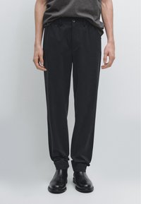 Sorte bukser med lige snit, lavet i glat stof, med frontlommer og knaplukning. Båret med sorte ankelstøvler.