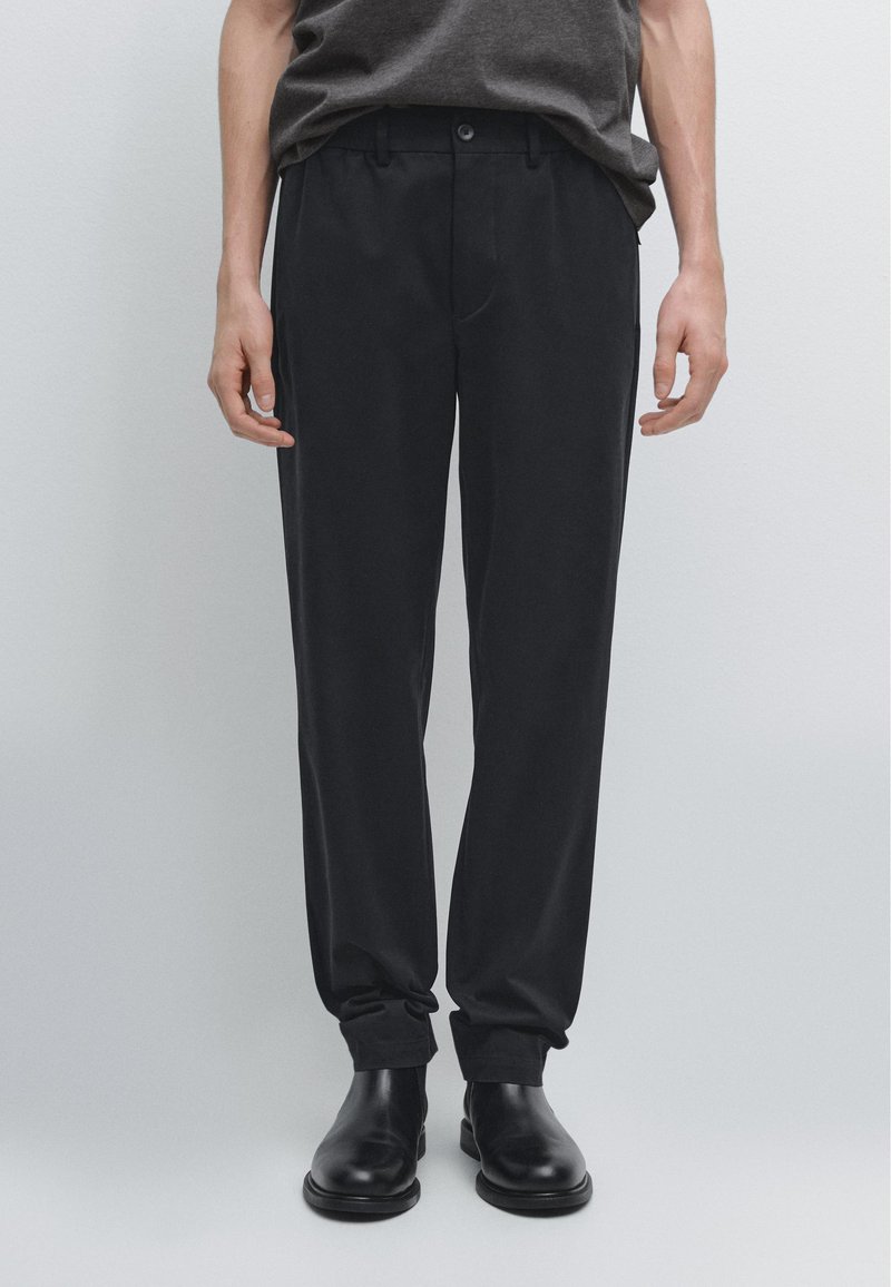 Sorte bukser med lige snit, lavet i glat stof, med frontlommer og knaplukning. Båret med sorte ankelstøvler.