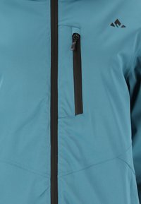 Türkise Jacke mit einem vorderen Reißverschluss mit schwarzen Akzenten, einer Brusttasche mit Reißverschluss und glatter, leichter Stoffstruktur.