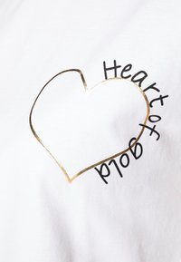 Bílá bavlněná trička s kovovým zlatým konturovým srdcem a textem "Heart of Gold" černou barvou, zakřiveným kolem tvaru srdce.