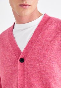 Detailní záběr na osobu, která má na sobě jasně růžový cardigan na knoflíky přes bílý tričko s kulatým výstřihem, zobrazuje dolní část obličeje a krk.