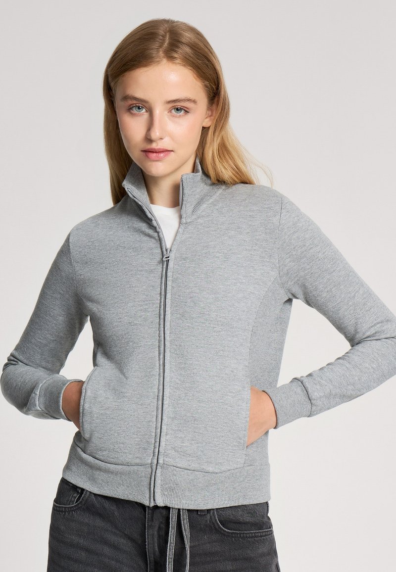 Felpa zip con colletto alto di colore grigio chiaro, con maniche lunghe, polsini a coste e tasche laterali, realizzata in tessuto morbido e texturizzato.