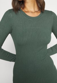 Žena oblečená v dlouhém rukávu, žebrovaných, tmavě zelených upnutých šatech s kulatým výstřihem a jemným zlatým řetízkem, s rukama opřenýma o boky.