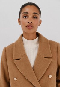 Cappotto doppiopetto color cammello in tessuto morbido, con ampi rever e due bottoni decorativi dorati. Indossato sopra un maglione bianco.