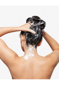 Person med vådt, mørkt hår, der påfører hvid shampoo på hovedbunden, set bagfra. Huden er glat uden synligt tøj.