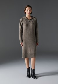 Robe pull en gris-brun, dotée d'une capuche et de détails côtelés, associée à des bottines noires à la cheville. Texture lisse, design mi-long.