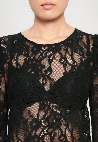 Gros plan sur une personne portant un haut en dentelle noire transparente avec des motifs floraux et un col rond.