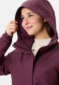 Giacca impermeabile bordeaux con zip frontale, cappuccio regolabile e polsini elastici. Presenta un logo sottile sul petto e una texture liscia.