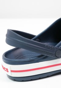 Gros plan d’un sabot Crocs bleu marine avec une semelle blanche comportant une bande rouge et une bride de talon réglable.